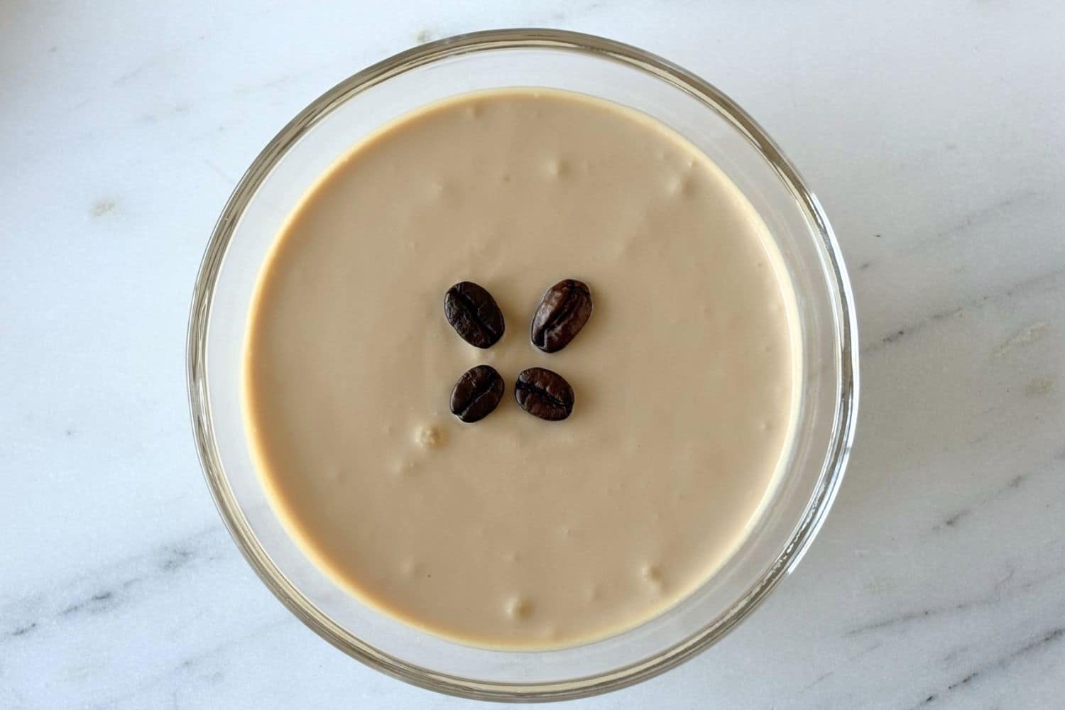 Top-down view of Marcella Hazan’s ricotta and coffee cream dessert in a glass bowl topped with four coffee beans on a marble surface.