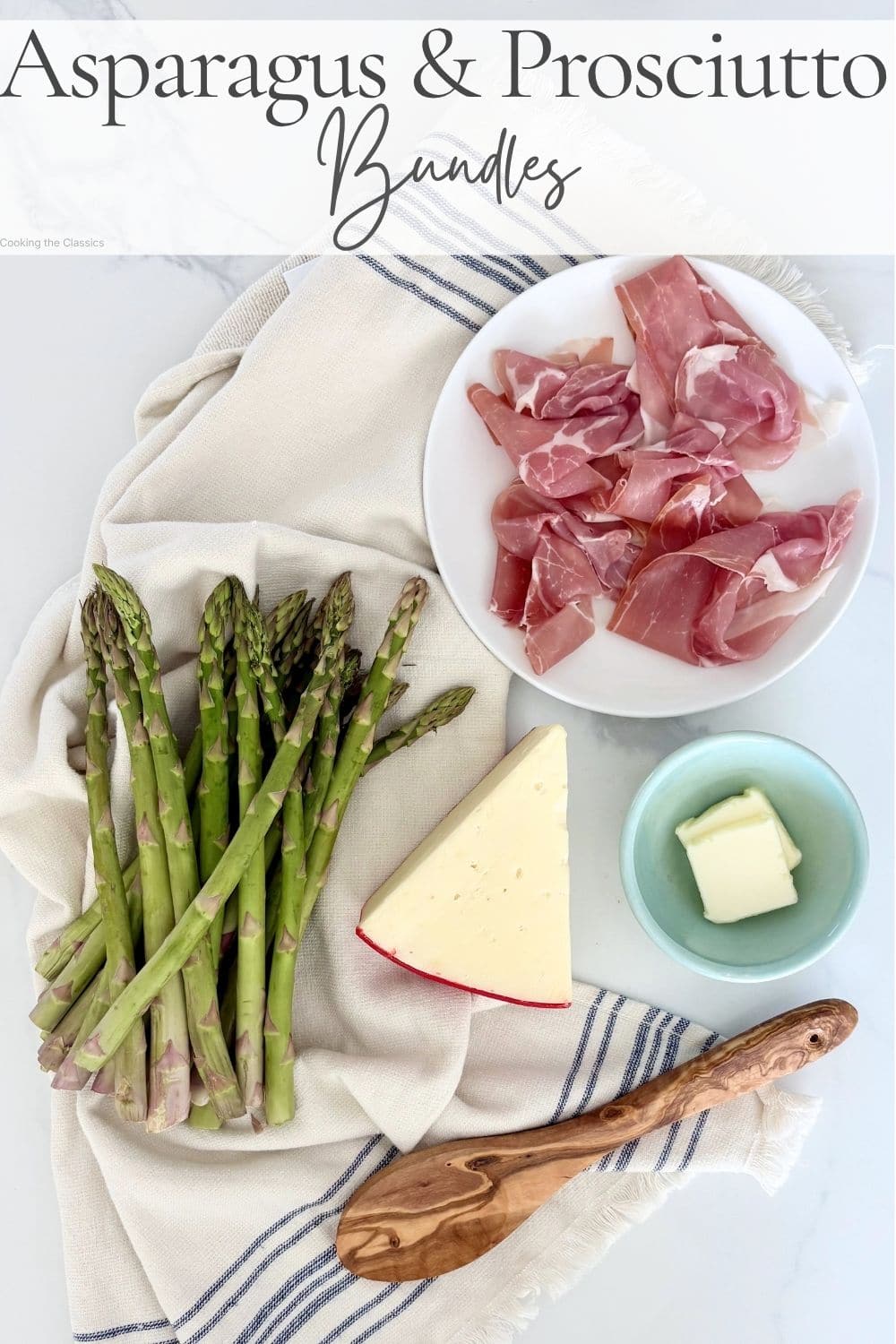 Flatlay of asparagus, prosciutto, cheese, and butter arranged on a marble surface with a linen and wooden spoon
