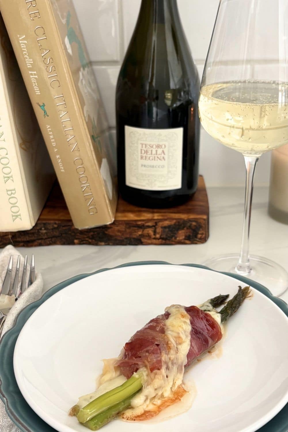 Asparagus and prosciutto bundles served with melted cheese on a plate, styled with prosecco and cookbooks