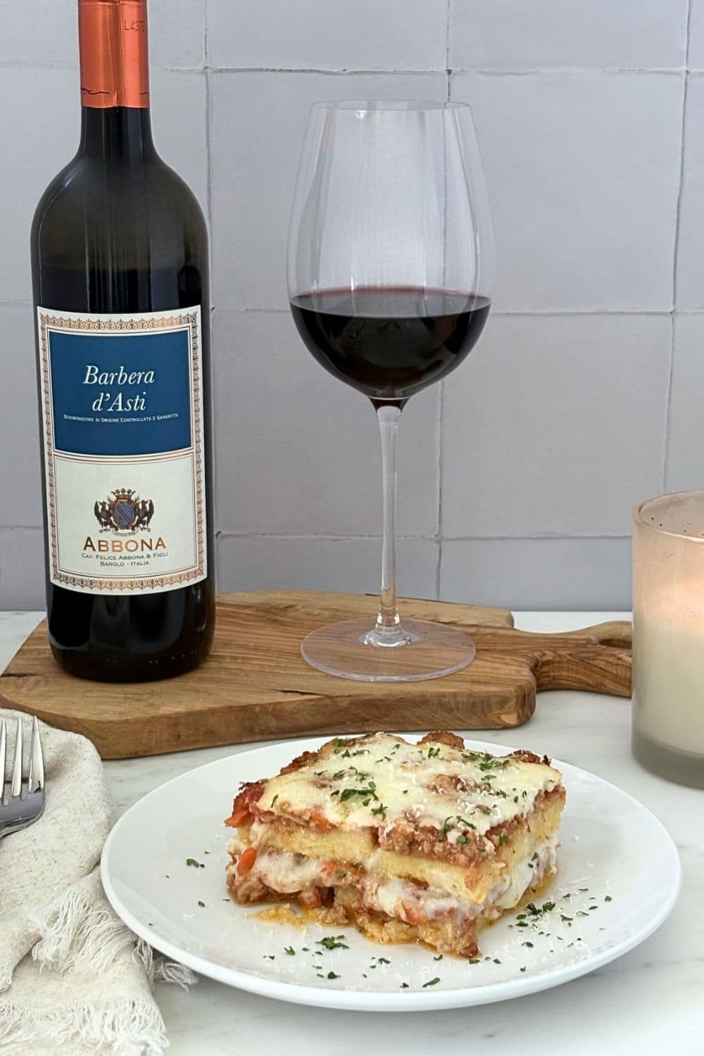 Slice of baked polenta with Bolognese and béchamel paired with a glass and bottle of Barbera wine on an olive wood board beside a candle.