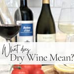 Two wine bottles with red and white wine glasses beside them, styled with tomatoes, lemons, and corks on a wooden board. Text overlay says What Does Dry Wine Mean?
