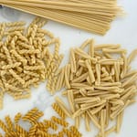 Assorted dried pasta shapes including spaghetti, fusilli, casarecce, and penne arranged on a white marble surface.