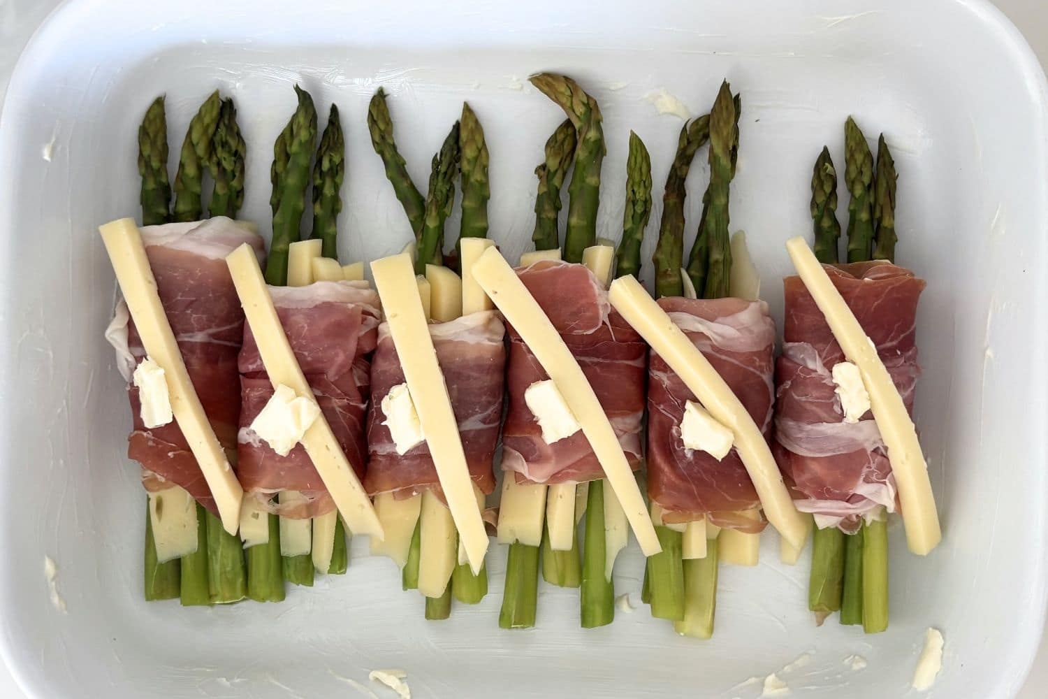 Asparagus bundles wrapped in prosciutto with slices of cheese and butter arranged in a baking dish before cooking