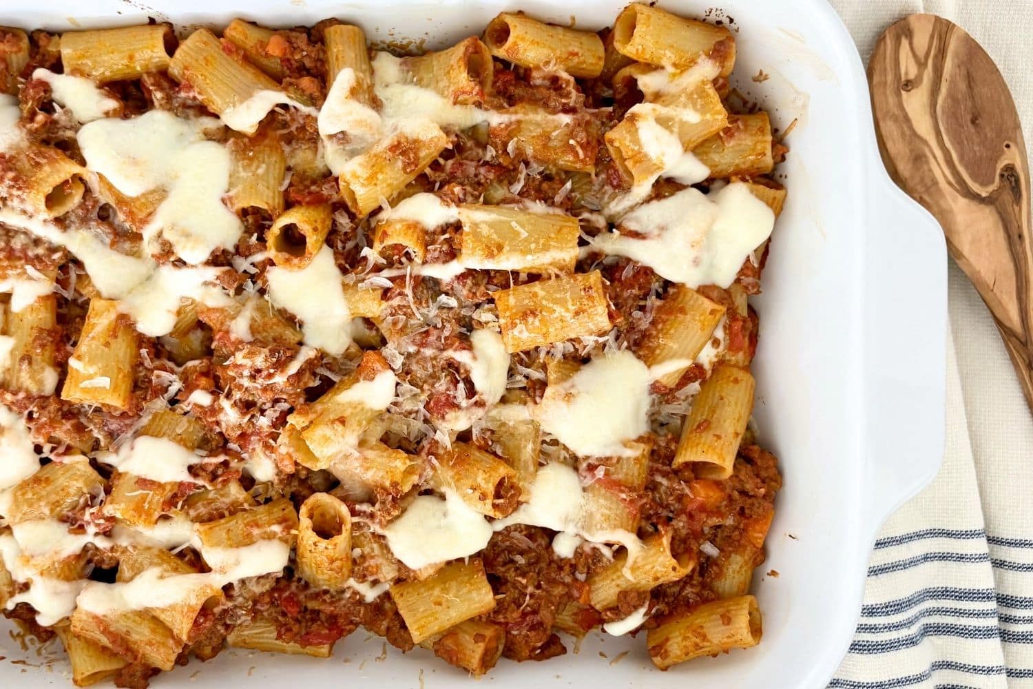 Baked rigatoni with Bolognese and béchamel in a white baking dish, topped with melted cheese and grated Parmesan, with a wooden spoon alongside.
