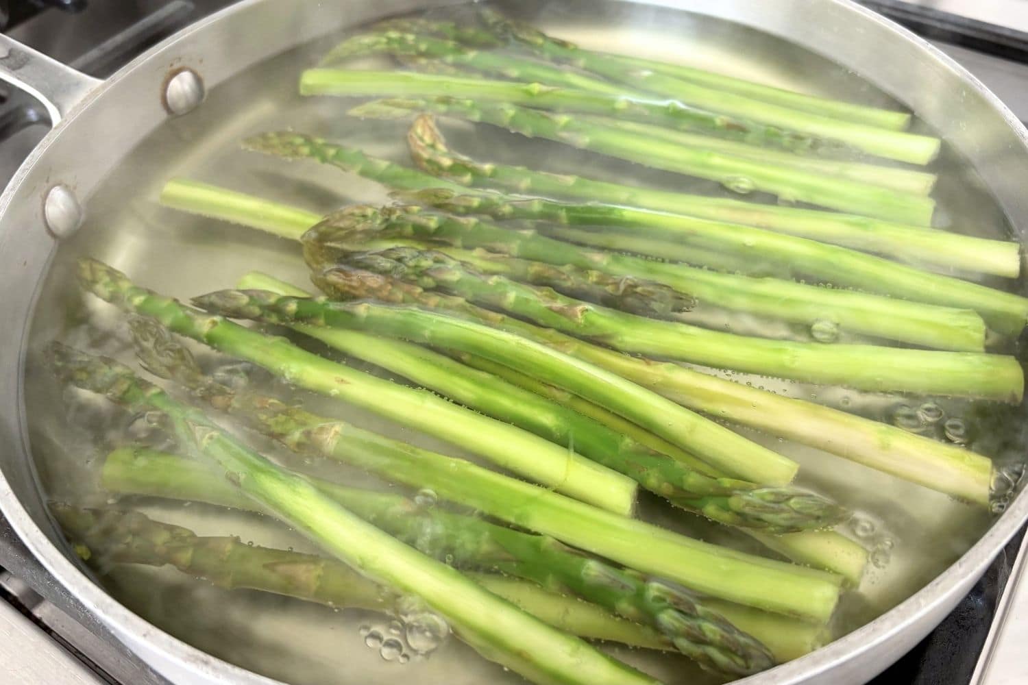 Blanching asparagus in a sauté pan of simmering water, showing bright green stalks cooking until just tender