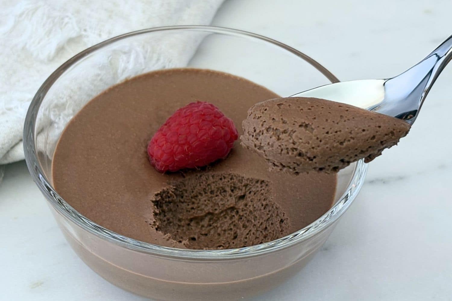 Chocolate mousse in a glass dessert bowl topped with a fresh raspberry, with a spoon lifting a light, airy bite to show the mousse's smooth texture.