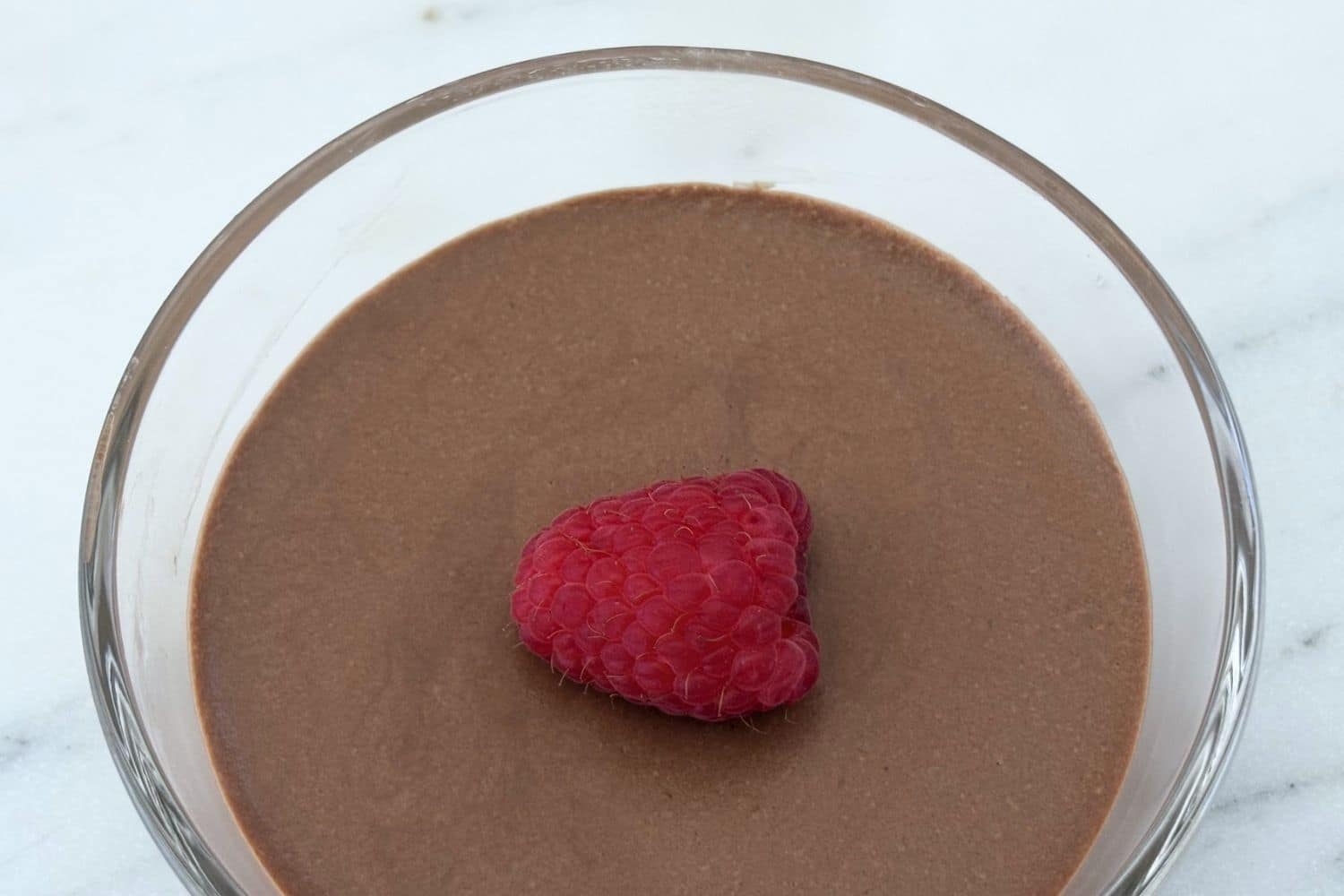 Overhead view of Italian chocolate mousse in a glass dessert bowl garnished with a single fresh raspberry on a smooth chocolate surface.