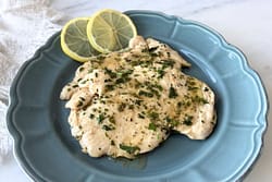 Thin chicken breast fillets with lemon parsley sauce served on a blue scalloped plate with lemon slices on a marble surface.