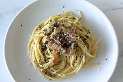 White bowl of Marcella Hazan’s carbonara sauce served with spaghetti, pancetta, parsley, black pepper, and grated cheese on a marble surface.