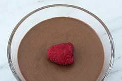 Overhead view of Italian chocolate mousse in a glass dessert bowl garnished with a single fresh raspberry on a smooth chocolate surface.