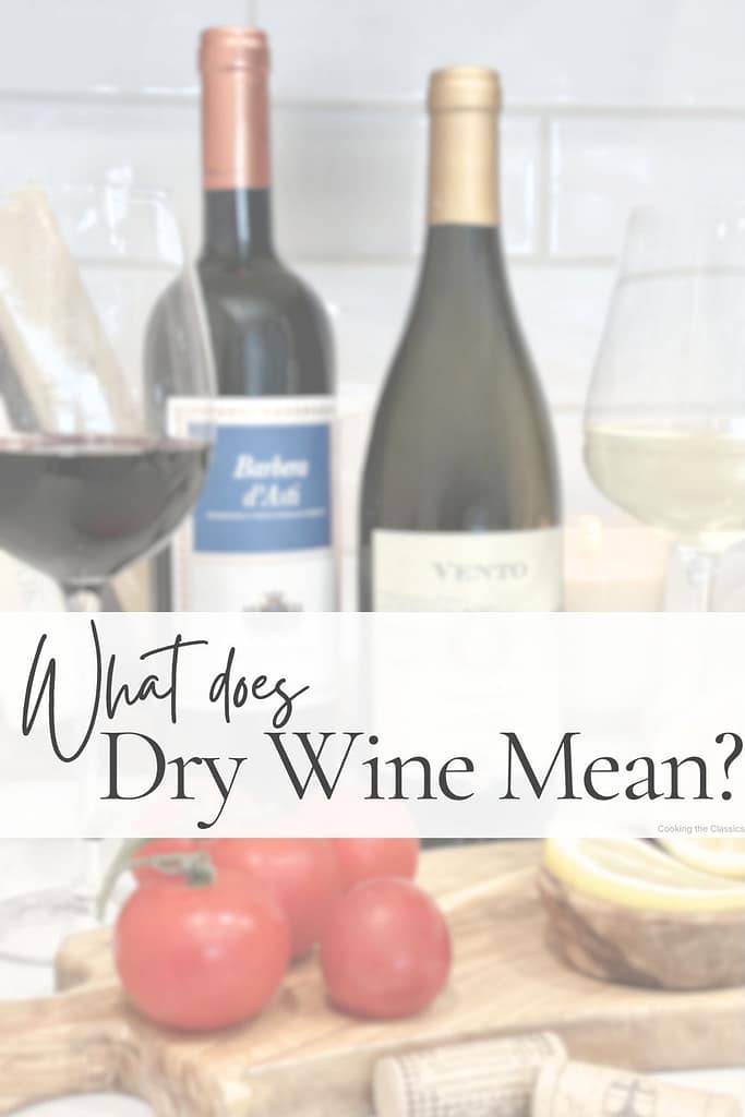 Two wine bottles with red and white wine glasses beside them, styled with tomatoes, lemons, and corks on a wooden board. Text overlay says What Does Dry Wine Mean?